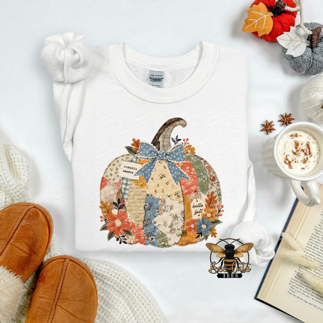 A white long-sleeve t-shirt with a colorful autumn patchwork pumpkin graphic, styled in a cozy flat-lay with a mug of hot cocoa, an open book, knit socks, and tan slippers.