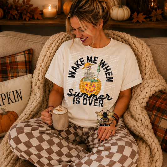 A woman sitting on a couch wrapped in a chunky knit blanket, wearing a white graphic t-shirt that reads 'WAKE ME WHEN Summer IS OVER' with a skeleton and pumpkin illustration, paired with brown and white checkered pajama pants.