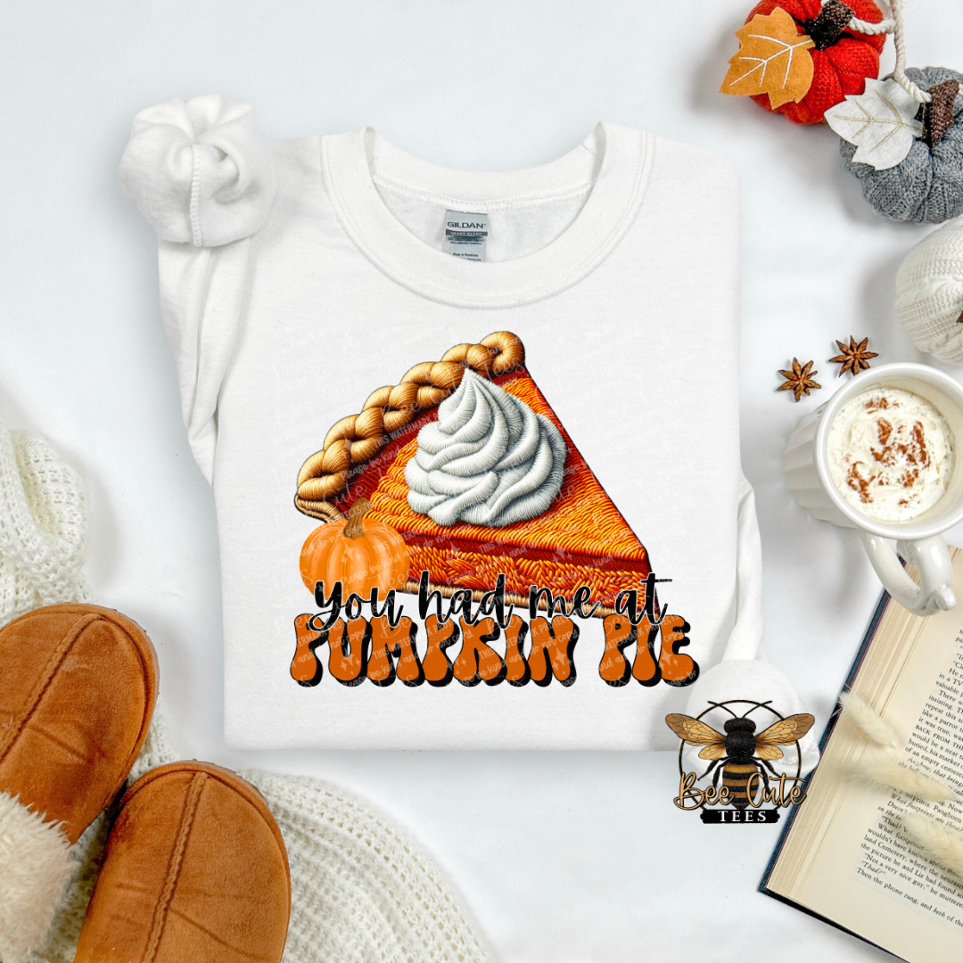 A white crewneck sweatshirt laid flat on a light background, featuring a graphic of an orange pumpkin pie slice with whipped cream and the text 'you had me at PUMKIN PIE' in orange and black. The sweatshirt is styled with autumn props including a mug of coffee, an open book, knit socks, and a small decorative bee logo watermark.