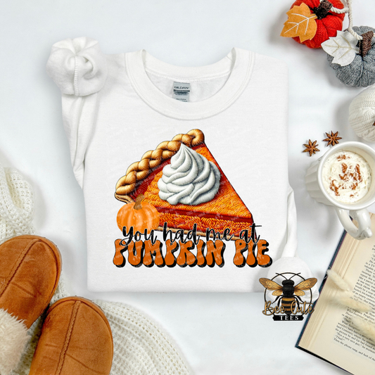 A white crewneck sweatshirt laid flat on a light background, featuring a graphic of an orange pumpkin pie slice with whipped cream and the text 'you had me at PUMKIN PIE' in orange and black. The sweatshirt is styled with autumn props including a mug of coffee, an open book, knit socks, and a small decorative bee logo watermark.