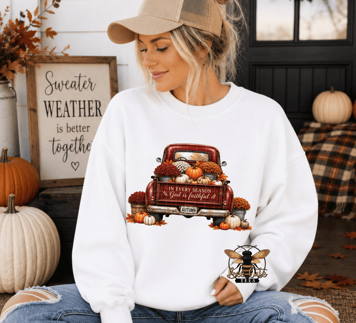 A woman wearing a white crewneck sweatshirt with a fall-themed graphic of a red pickup truck loaded with pumpkins and autumn decorations. The sweatshirt features a bee logo on the bottom right.