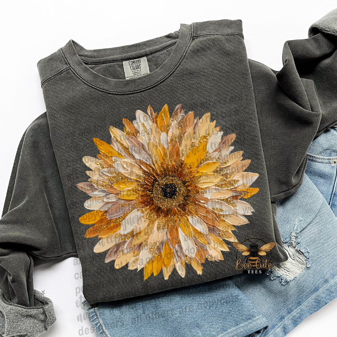 A dark charcoal gray sweatshirt featuring a large, textured sunflower graphic on the front in shades of yellow, orange, and brown. The sweatshirt is styled in a flat-lay arrangement with light blue distressed denim jeans on a white background.