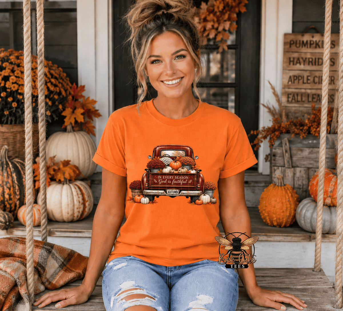 A woman sitting on a porch swing wearing an orange graphic t-shirt with a red pickup truck carrying pumpkins and autumn leaves. The shirt has a vintage truck graphic and a bee logo watermark in the lower right corner.