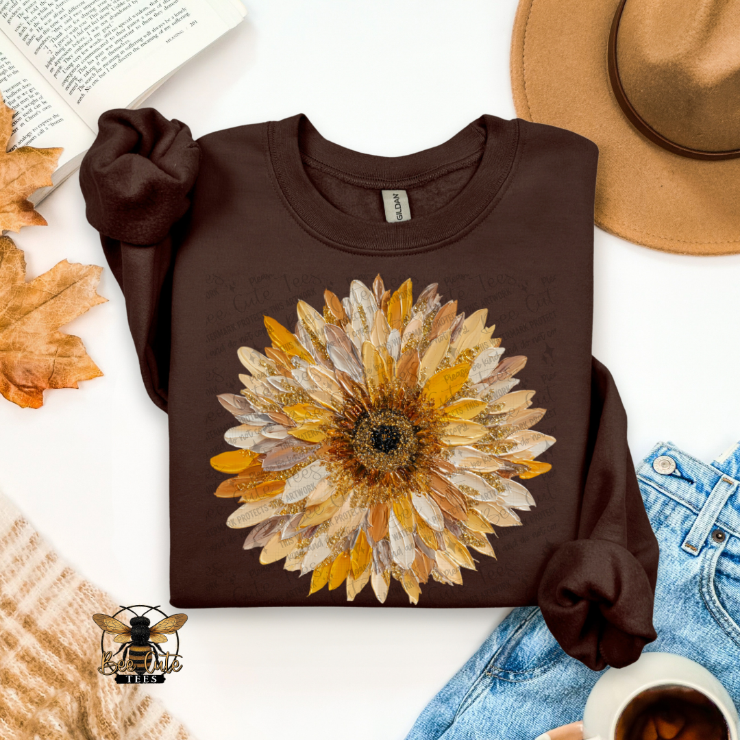 A dark brown long-sleeve t-shirt with a large autumn sunflower graphic in yellow, gold, and beige tones, styled in a flat-lay arrangement with a tan hat, blue jeans, a book, leaves, and a coffee cup.