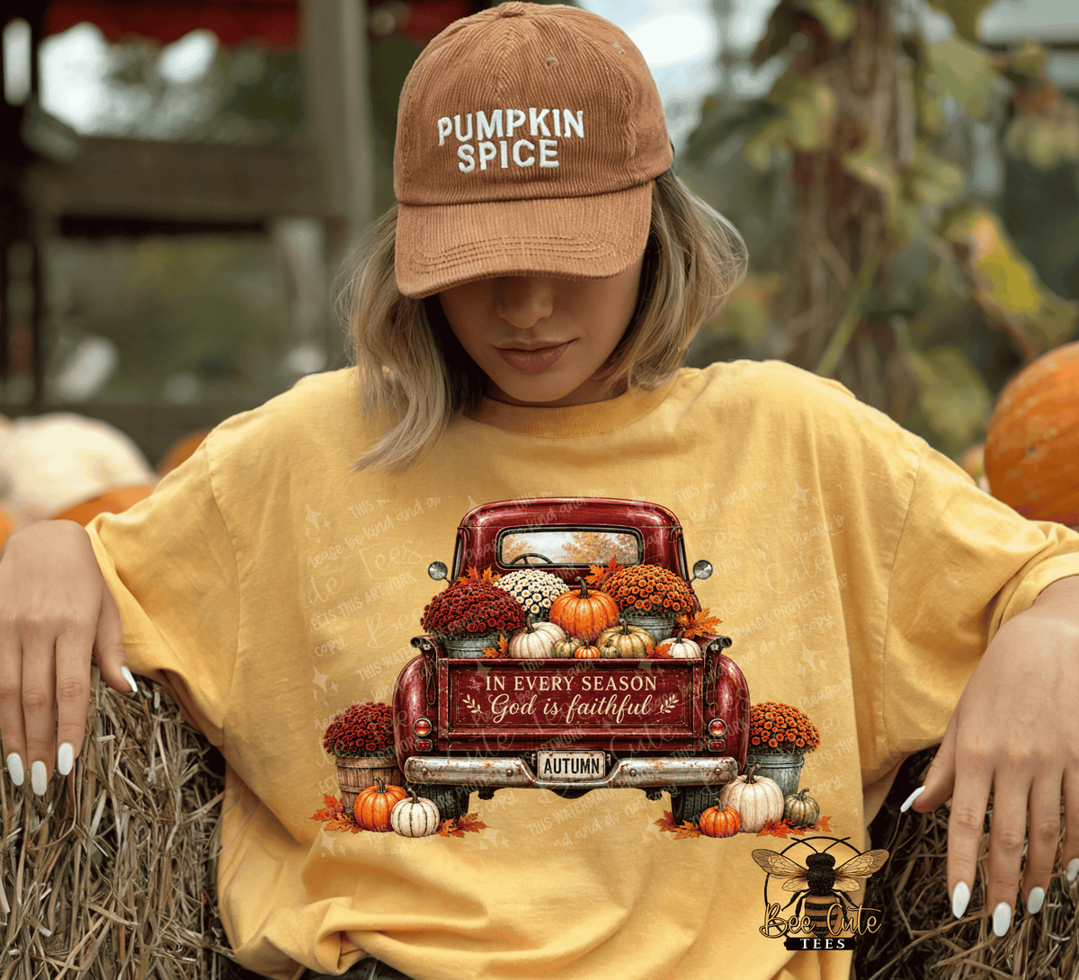 A woman wearing a mustard yellow graphic t-shirt and a brown 'PUMPKIN SPICE' dad hat, sitting on a hay bale at an outdoor autumn-themed event. The t-shirt features a graphic of a red pickup truck filled with pumpkins and fall foliage, with the text 'IN EVERY SEASON God is faithful' and 'AUTUMN'.