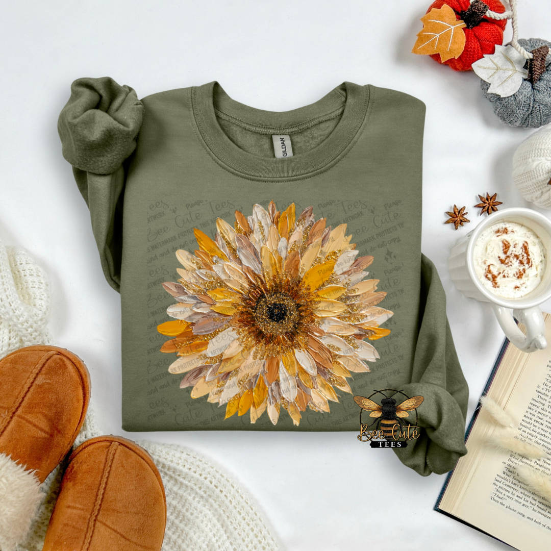 An olive green long-sleeve sweatshirt with a large autumn sunflower graphic on the front, styled in a cozy flat-lay with brown slippers, a mug of coffee, an open book, and fall leaves.