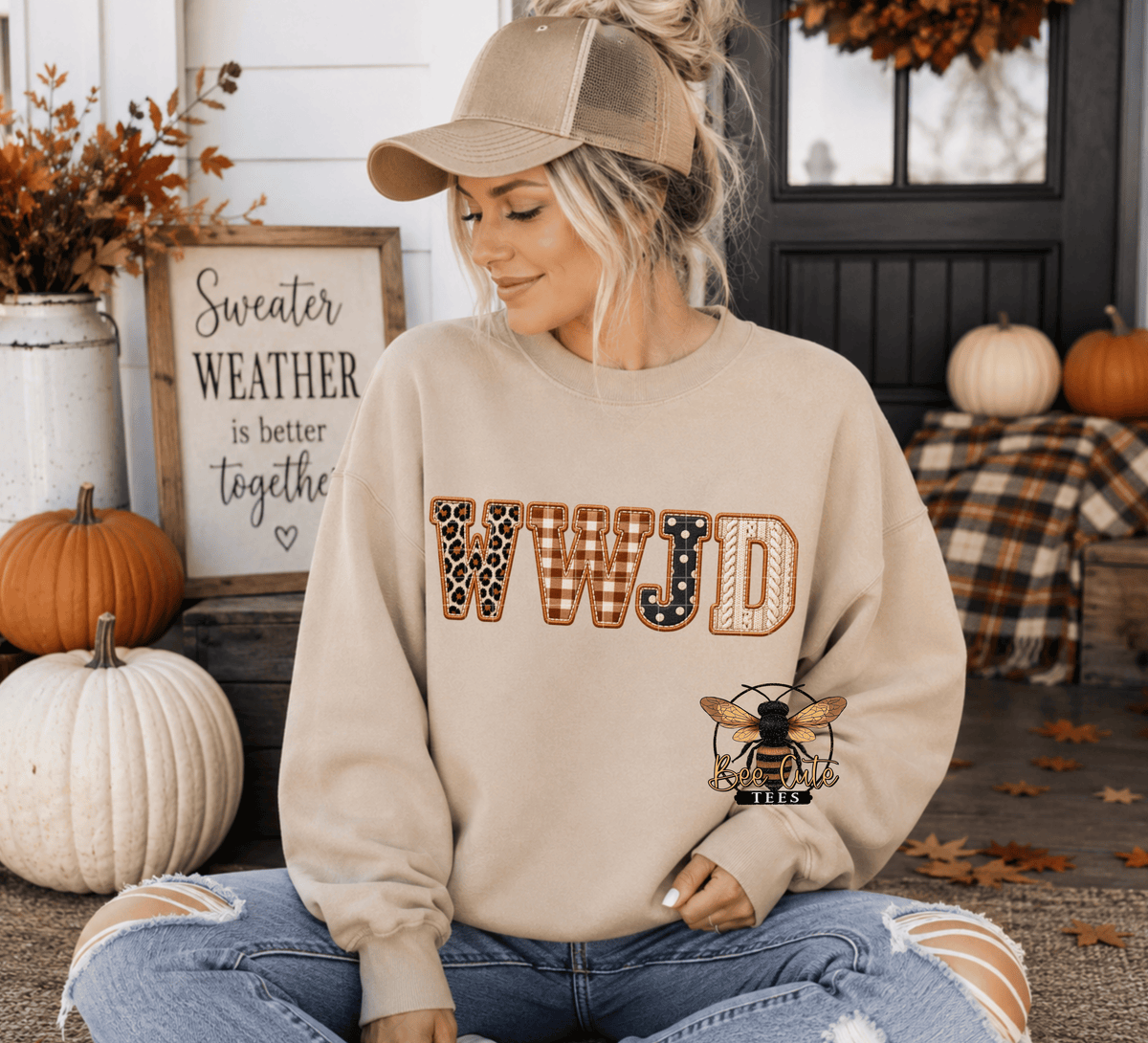 A woman wearing a beige crewneck sweatshirt with 'WWJD' in a plaid and leopard print pattern, and a bee logo on the lower right. She is sitting on a porch decorated with pumpkins and autumn leaves.
