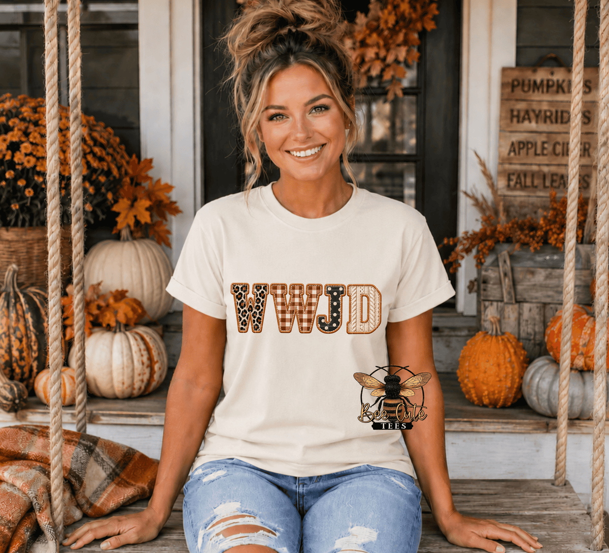 A woman sitting on a porch swing wearing a cream-colored t-shirt with 'WWJD' printed in a leopard and patterned font, paired with distressed blue jeans. The background features autumn decorations including pumpkins, fall foliage, and a rustic wooden sign.