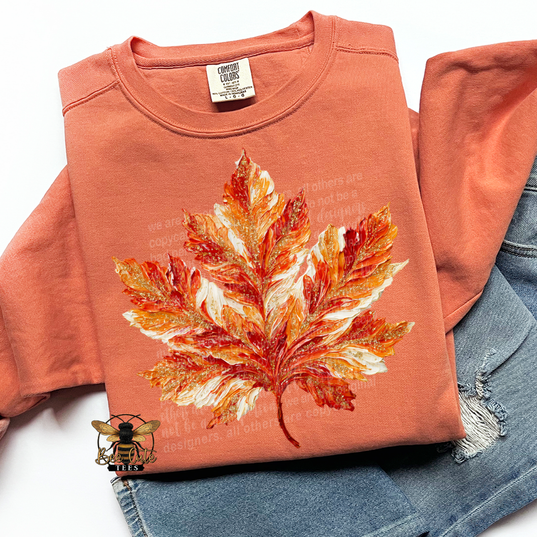 A folded terracotta orange long-sleeve t-shirt with a large autumn maple leaf graphic in orange, red, and white on the front, styled flat-lay with blue denim jeans underneath. 