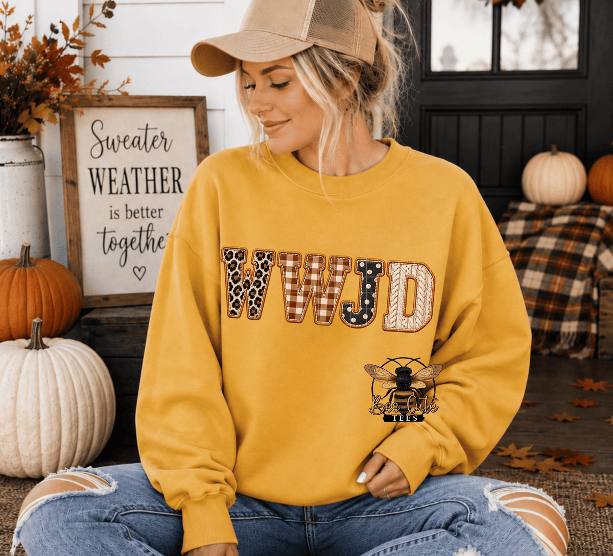 A woman wearing a mustard yellow sweatshirt with 'WWJD' in a fall-themed patchwork design and a bee logo on the front, sitting in a decorated autumn porch setting.
