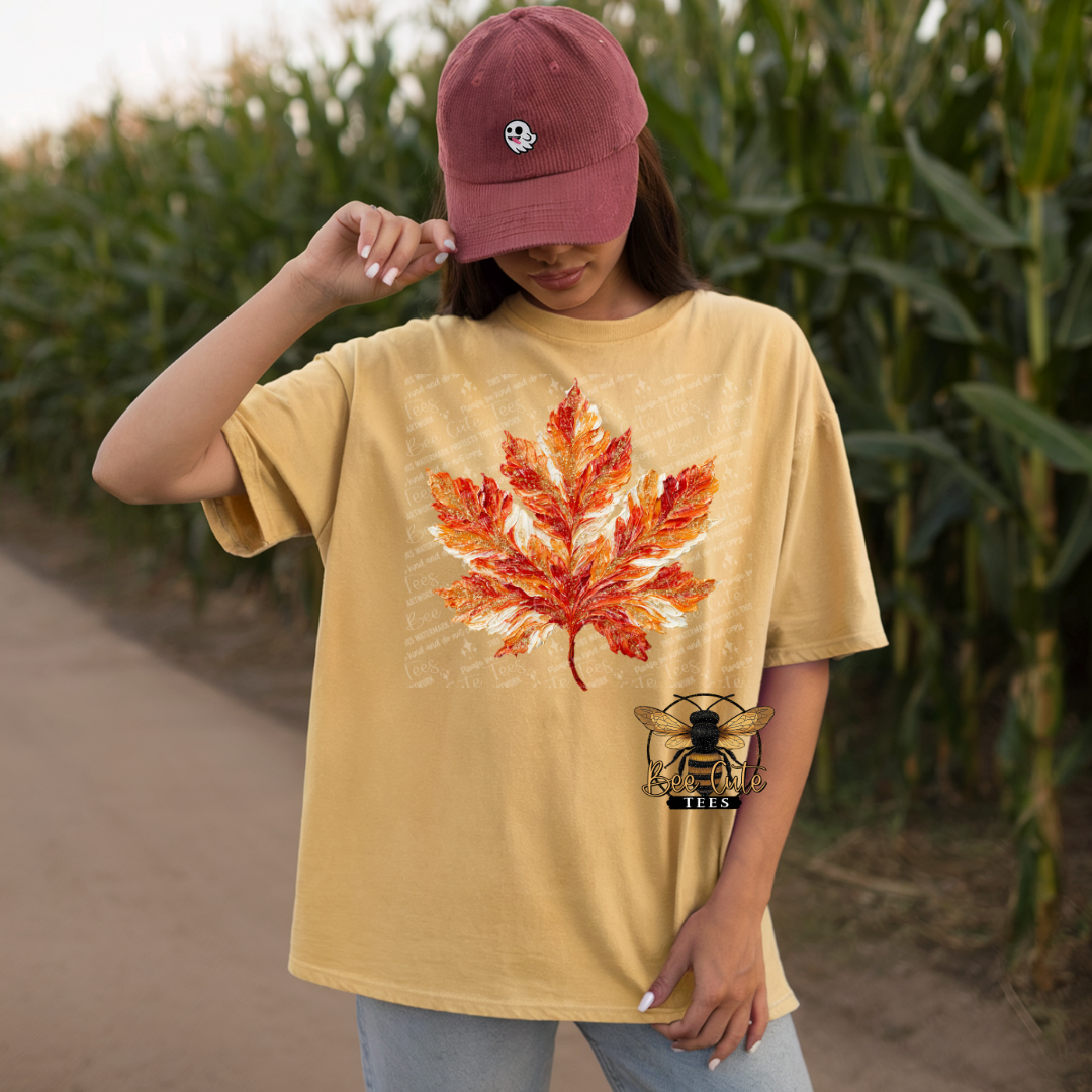 A woman wearing a tan oversized graphic t-shirt with a large orange and red maple leaf print on the front, a maroon baseball cap, and light blue jeans, standing in a cornfield.