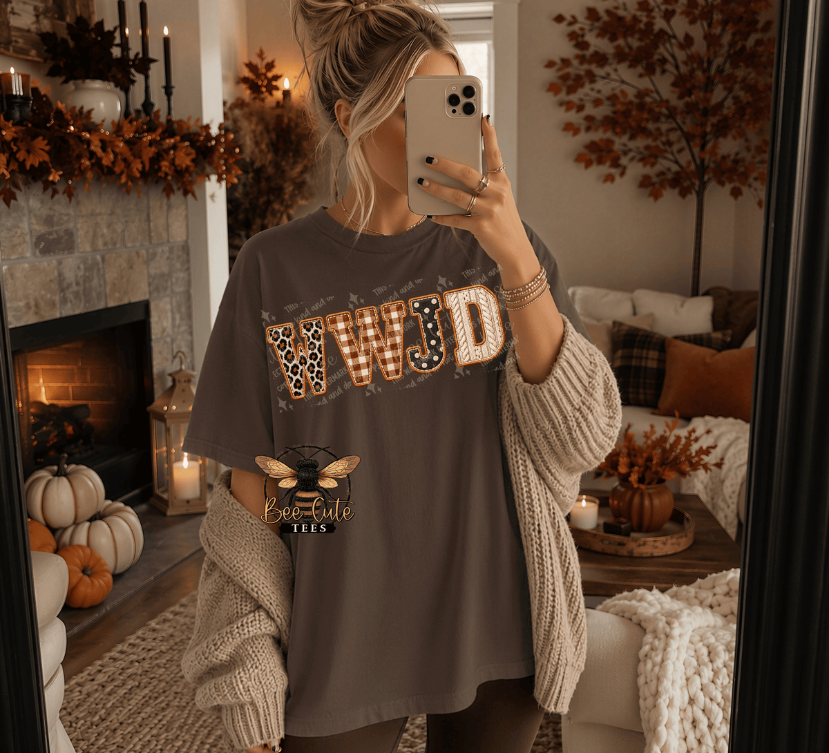 A woman takes a mirror selfie in a cozy living room, wearing a dark brown graphic t-shirt with 'WWJD' in leopard and striped lettering, a chunky cream knit cardigan, and dark leggings. The shirt features a bee logo with the text 'Be Cute TEEs'.