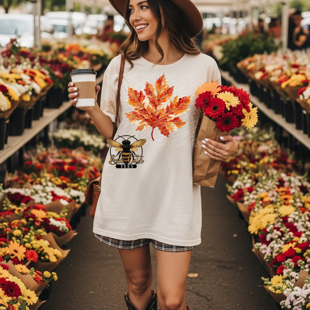 A woman in a flower market aisle wearing a cream-colored oversized t-shirt with a large orange and red maple leaf graphic and a bee design on the front, holding a coffee cup and a paper bag of flowers.