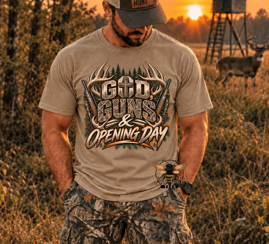 A man wearing a tan graphic t-shirt with 'God, Guns & Opening Day' printed on the front, standing in a grassy field at sunset with a deer and hunting stand in the background.