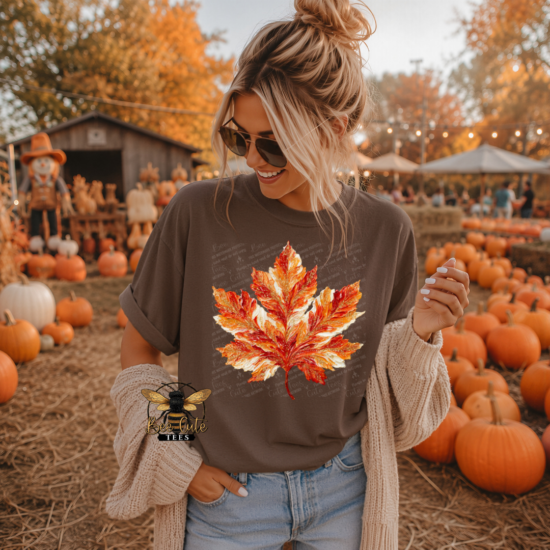 A woman wearing a brown graphic t-shirt with a large red, orange, and yellow maple leaf print, styled with a beige knit cardigan and sunglasses, standing in a pumpkin patch.