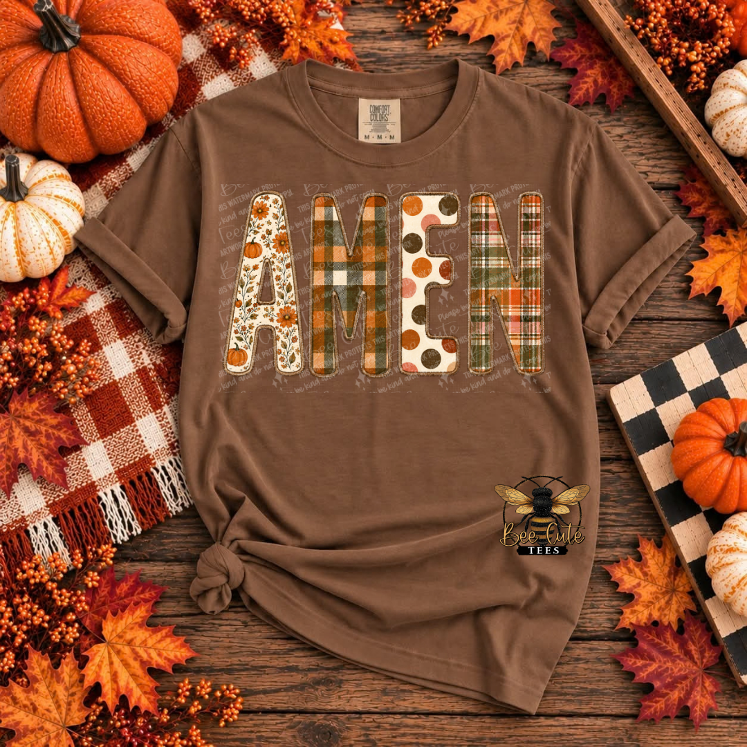 A brown short-sleeve graphic T-shirt laid flat on a rustic wood surface, surrounded by autumn decorations including pumpkins, fall leaves, berries, and a plaid blanket. The shirt has the word "AMEN" spelled out in decorative patterned letters and a small bee logo with the text "BEE CUTE TEES" near the lower right front.
