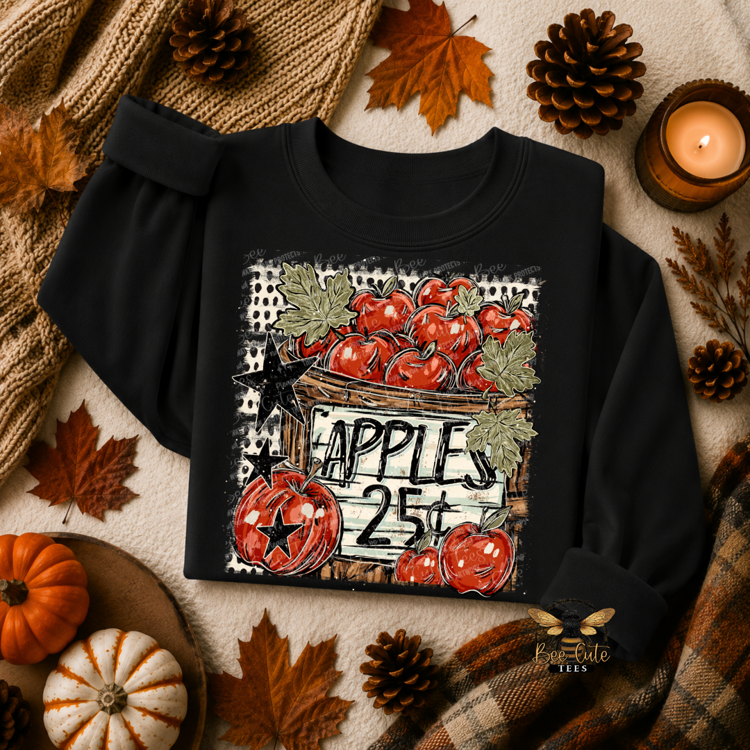 A black long-sleeve crewneck sweatshirt with a rustic autumn graphic of red apples and a pumpkin that reads 'APPLES 25¢'. The sweatshirt is styled in a cozy fall flat-lay with a knit blanket, plaid scarf, pumpkins, pinecones, and a lit candle.