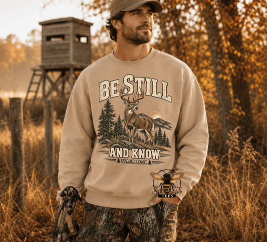 A man wearing a beige crewneck sweatshirt with a deer hunting graphic that reads 'BE STILL AND KNOW', standing outdoors in a wooded setting with a wooden blind in the background.