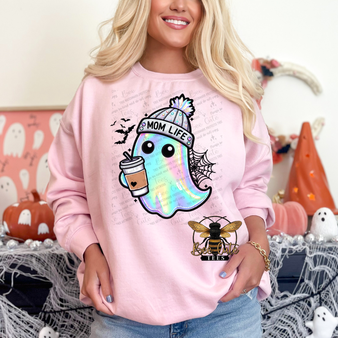 A woman wearing a light pink crewneck sweatshirt with a colorful graphic of a ghost holding an iced coffee and the text 'MOM LIFE' alongside a small bee logo. The background is decorated for Halloween with pumpkins, spiderwebs, and ghost decorations.