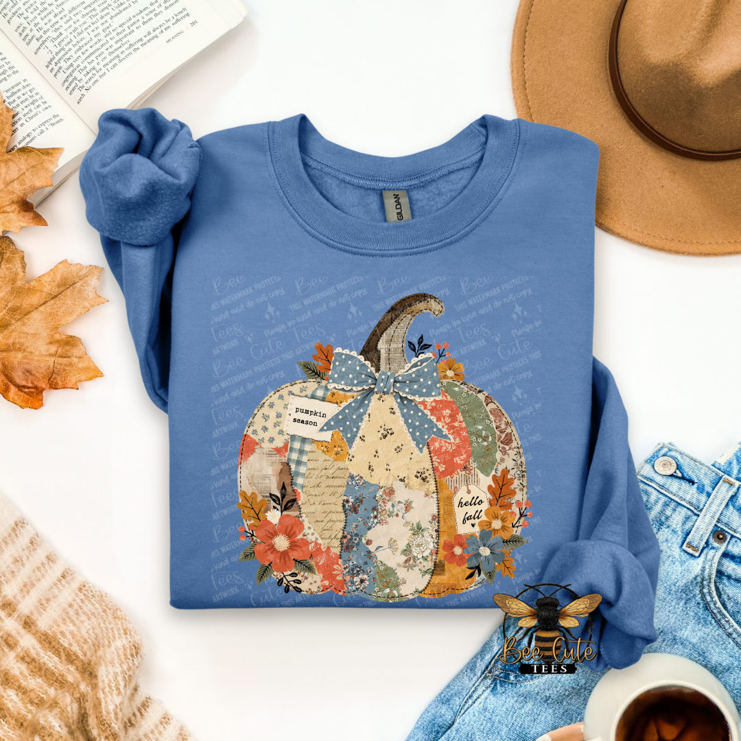 A folded blue long-sleeve crewneck sweatshirt with a colorful autumn pumpkin graphic, styled in a flat lay with a tan hat, denim jeans, a book, leaves, and a cup of coffee.