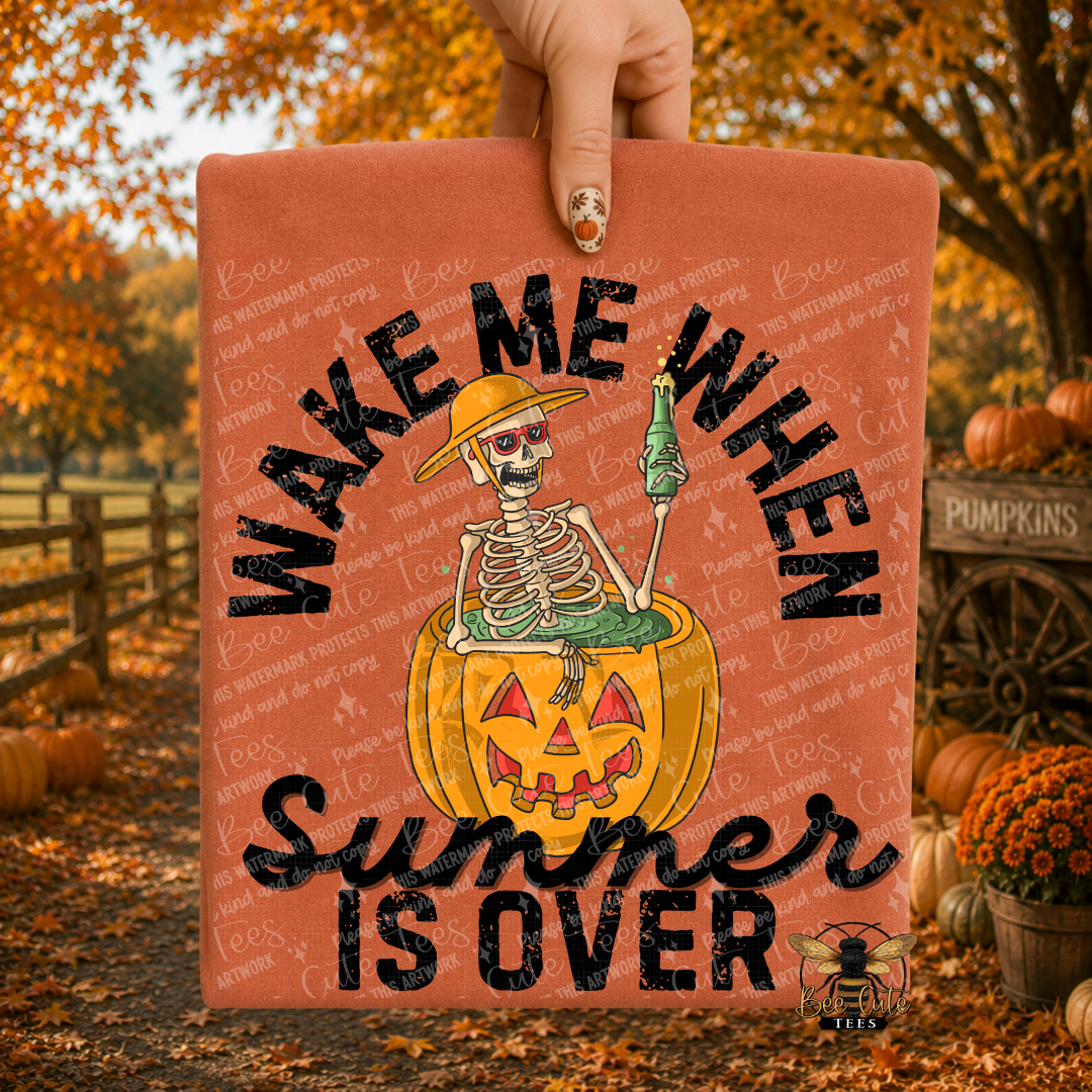 A hand holds up an orange-colored rectangular sign or print featuring a cartoon skeleton sitting in a jack-o'-lantern pumpkin, with the text 'WAKE ME WHEN Summer IS OVER' in black. The background shows an autumn outdoor scene with pumpkins, trees, and fallen leaves.