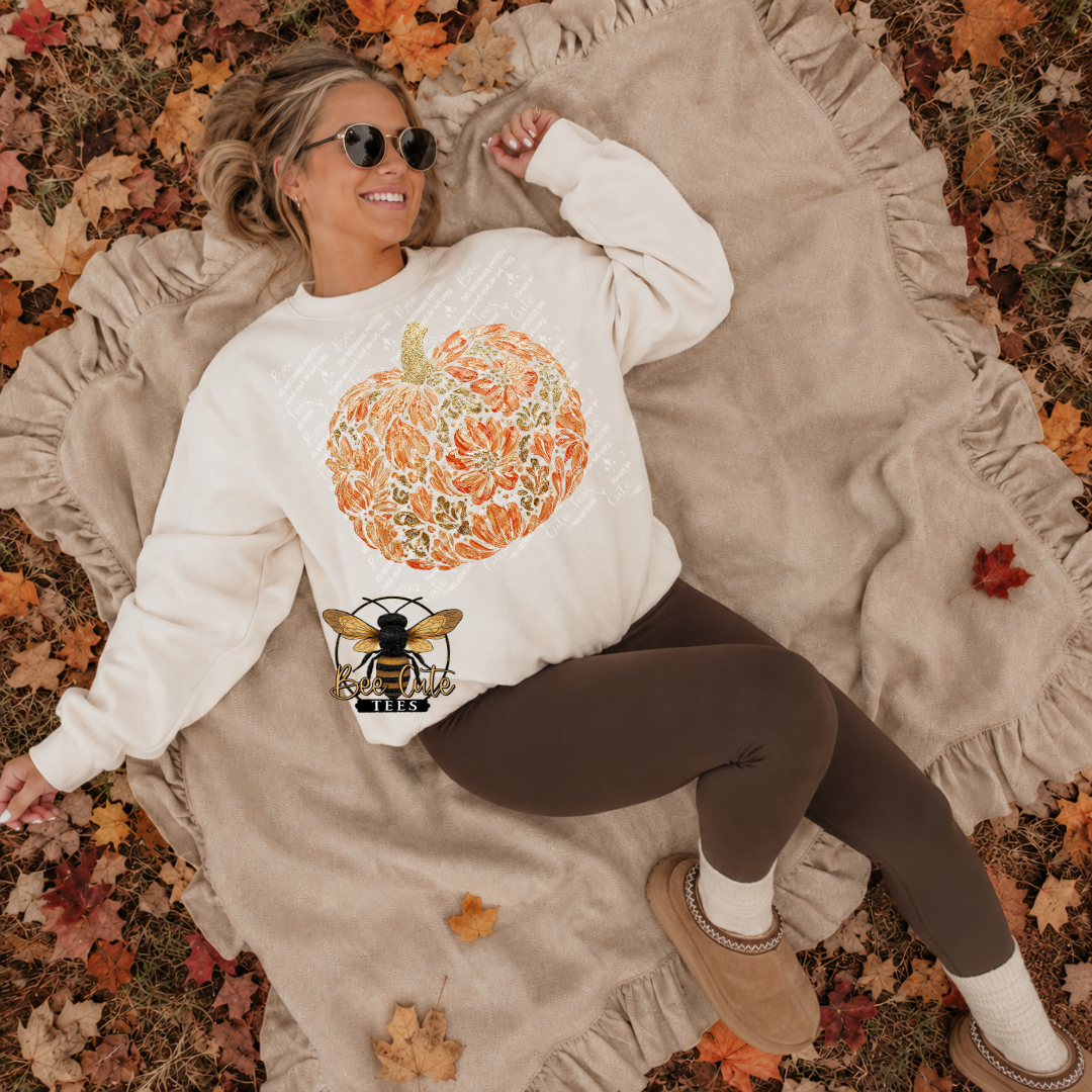 A woman lying on a beige blanket surrounded by fallen autumn leaves, wearing a cream sweatshirt with a large orange pumpkin graphic and a bee patch graphic, brown leggings, white socks, and tan slippers.
