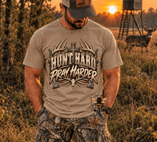 A man wearing a tan graphic t-shirt with 'HUNT HARD PRAY HARDER' printed on the front, standing in a grassy outdoor field at sunset with a deer and hunting stand in the background.