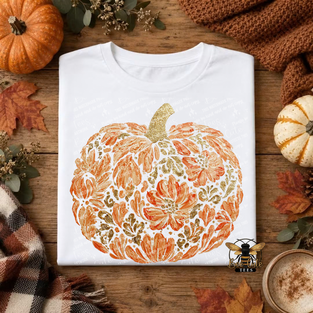 A folded white T-shirt with a large autumn pumpkin graphic in orange, gold, and brown tones, styled on a rustic wooden surface with fall leaves, a small pumpkin, a knit sweater, and a cup of coffee.