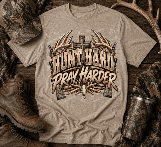 A beige graphic t-shirt laid flat on a rustic wood surface, surrounded by hunting-themed props including camouflage boots, a camo hat, antlers, and a camouflage cup. The shirt features a large front graphic with the words 'HUNT HARD PRAY HARDER' in distressed brown and tan lettering, flanked by antlers, a cross, and a skull motif.