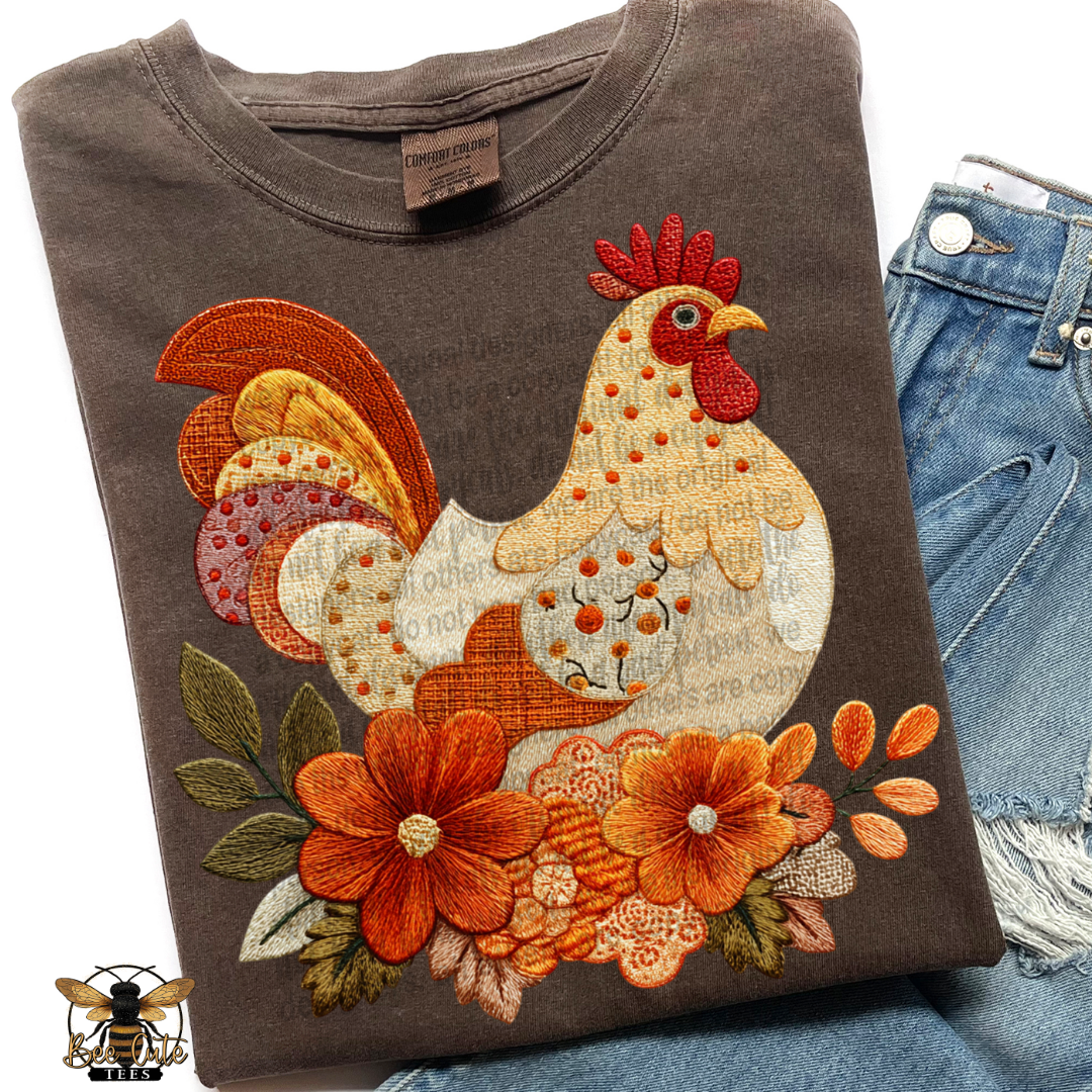 A folded dark brown t-shirt featuring a large, colorful patchwork-style embroidered rooster and floral design, styled next to a pair of blue denim jeans on a white background.