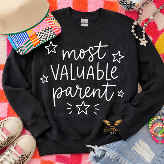 A black crewneck sweatshirt with the phrase 'most VALUABLE parent' in white script and star graphics, styled in a flat lay with a hat, sneakers, jeans, beads, and a disco ball on a pink and orange patterned blanket.