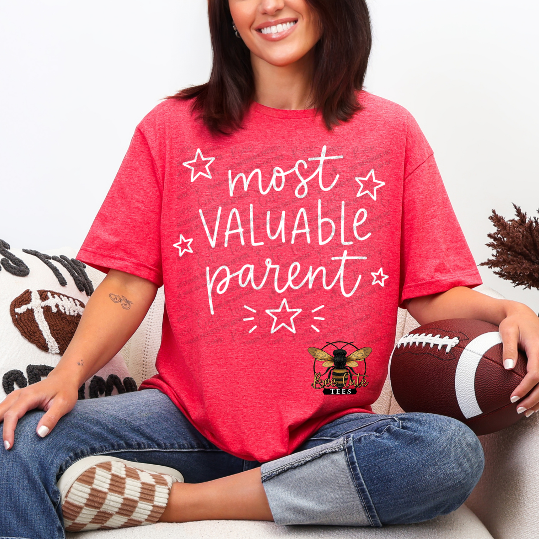 A woman sitting on a couch wearing a heather red graphic t-shirt that reads 'most VALUABLE parent' in white script with star and dash accents, along with a bee logo. She is holding a football.