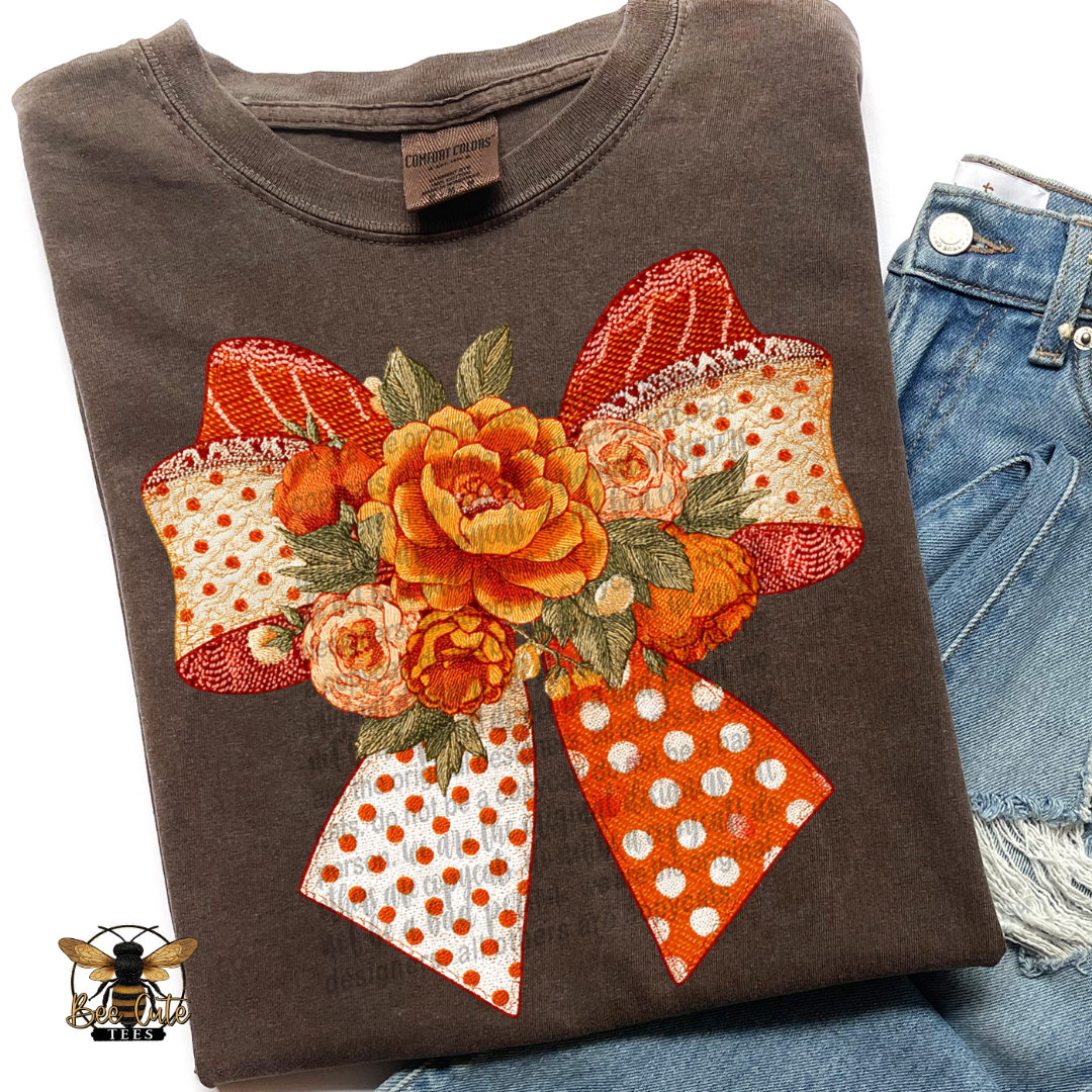 A folded dark brown t-shirt featuring a large orange and red floral bow graphic on the front, styled next to a pair of blue denim shorts.
