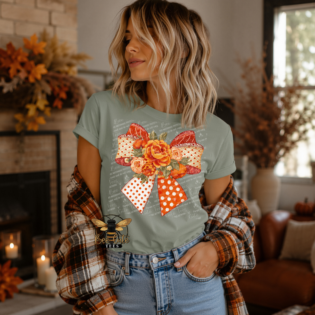 A woman wearing a sage green graphic t-shirt with a large orange and white polka dot bow and floral design, styled with a brown and orange plaid flannel shirt and blue jeans in a cozy, warmly lit living room.