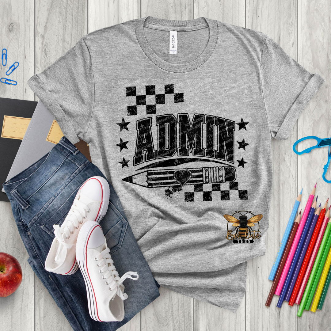 A heather gray graphic T-shirt laid flat on a light wood surface, styled with blue jeans, white sneakers, colored pencils, a red apple, and small stationery props. The shirt has a large black 'ADMIN' graphic with stars, checkered flags, a pencil illustration, and a bee logo near the lower right.