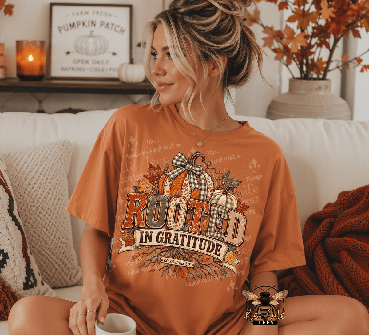 A woman sits on a white couch wearing a rust-orange graphic T-shirt with a fall harvest design that reads 'Rooted in Gratitude' and features pumpkins, leaves, and a bow. The scene is styled with cozy home decor, including a candle, framed sign, pillows, and a throw blanket.