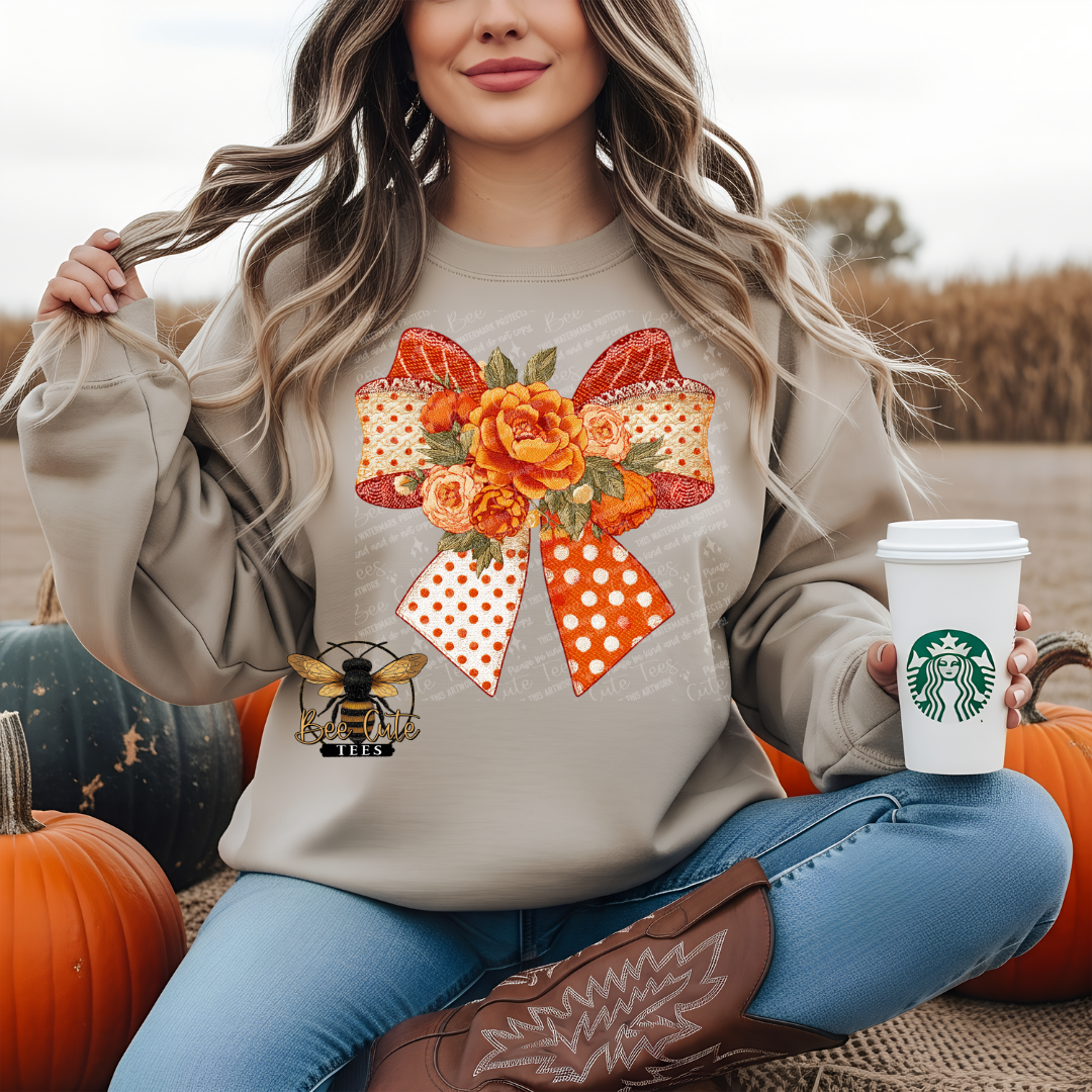 A woman sitting outdoors among pumpkins wears a beige sweatshirt with a large orange floral bow graphic on the front. She holds a white coffee cup in one hand and wears blue jeans and brown cowboy boots.