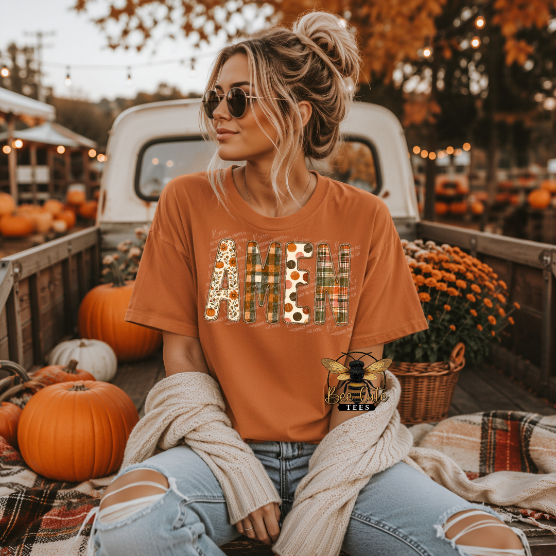 A woman wearing a burnt orange graphic t-shirt with 'AMEN' in autumn-themed patterns sits in the bed of a pickup truck decorated for fall, surrounded by pumpkins and mums.
