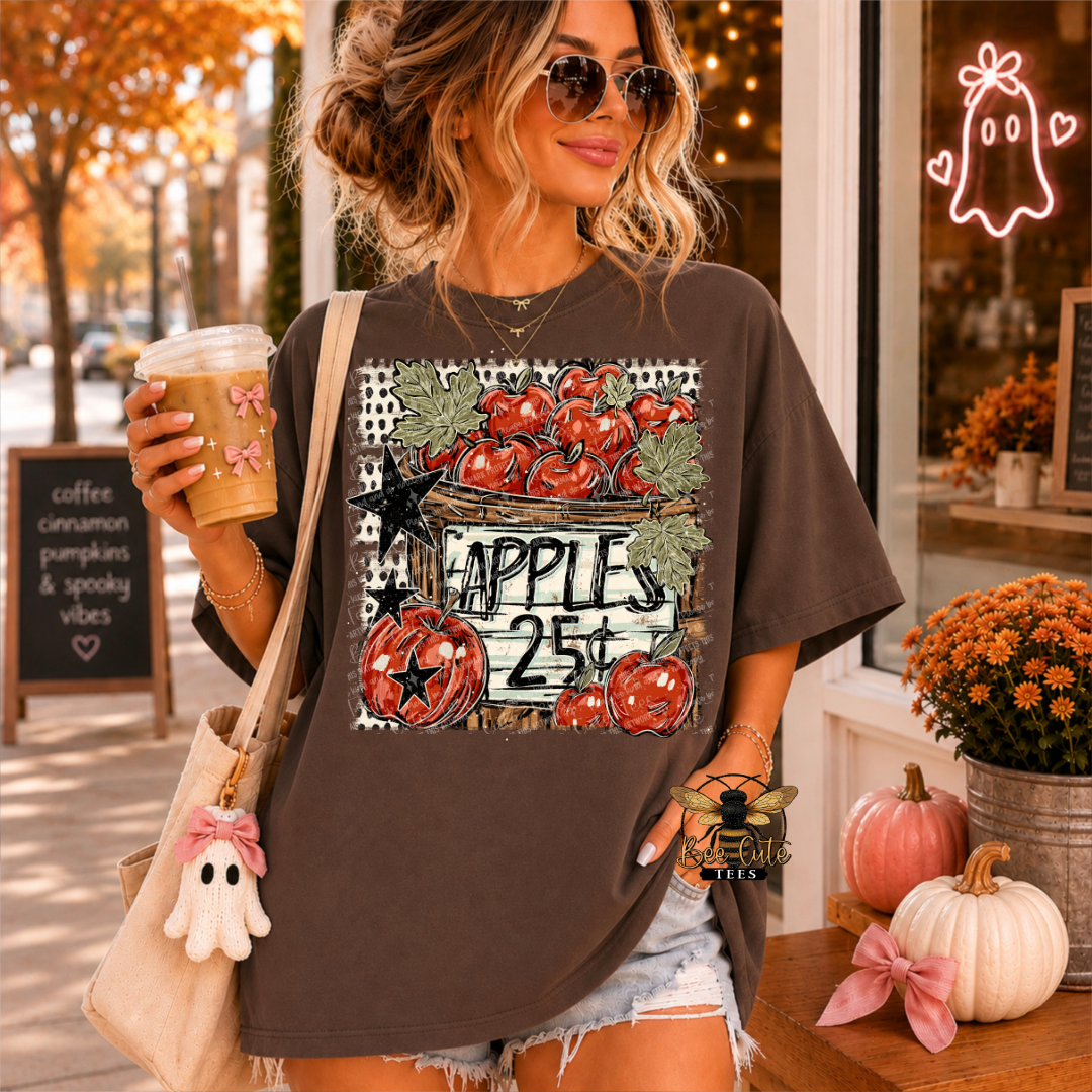 A woman wearing a dark brown graphic t-shirt with a Halloween apple design, holding a coffee cup and a tote bag in a fall-themed storefront setting.