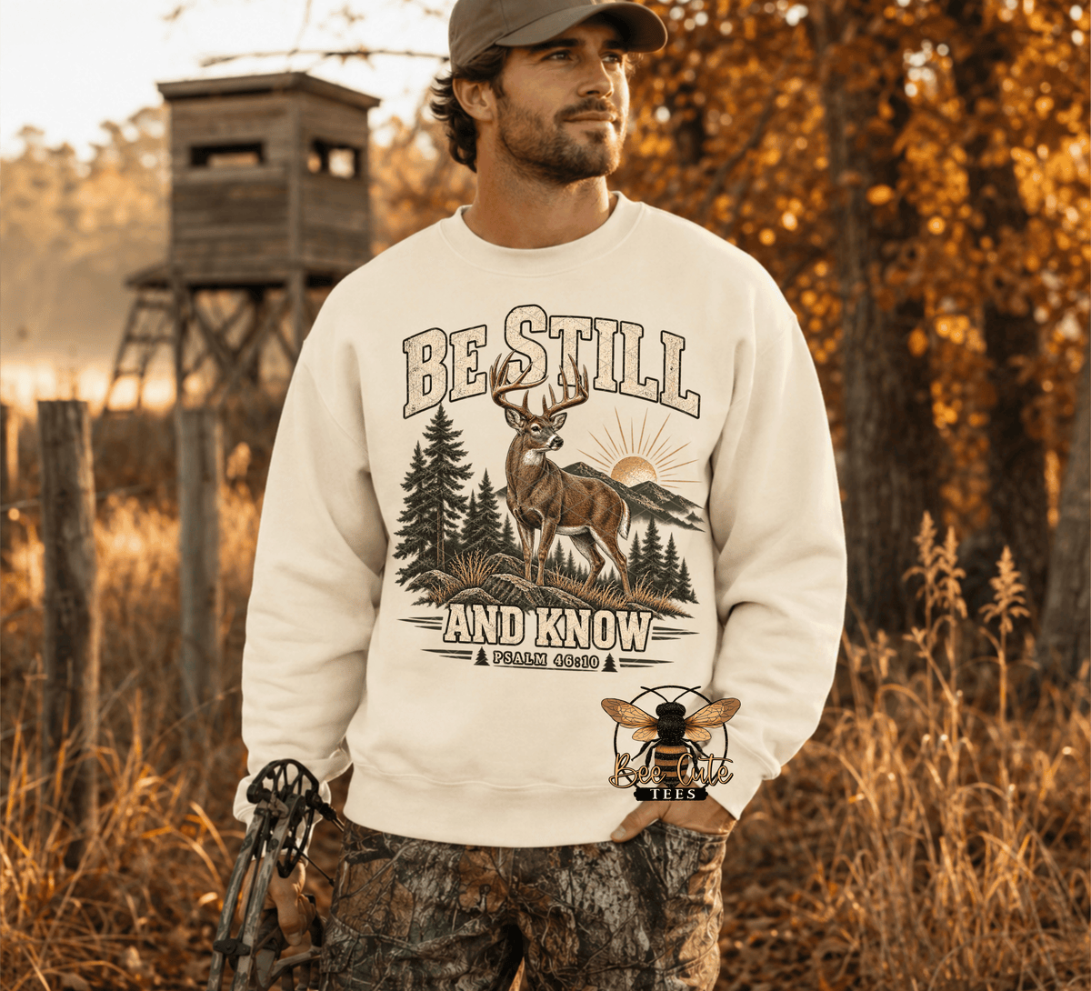 A man wearing a off white crewneck sweatshirt with a deer hunting graphic that reads 'BE STILL AND KNOW', standing outdoors in a wooded setting with a wooden blind in the background.
