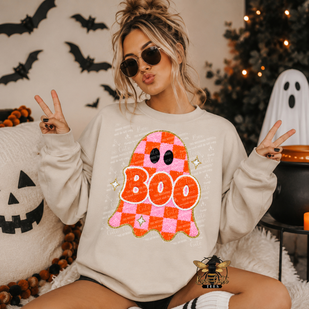 A woman wearing a cream-colored sweatshirt with a large pink and orange checkered ghost graphic that says 'BOO' in orange letters, posed in a cozy Halloween-themed room with pumpkins, bats, and a ghost decoration.