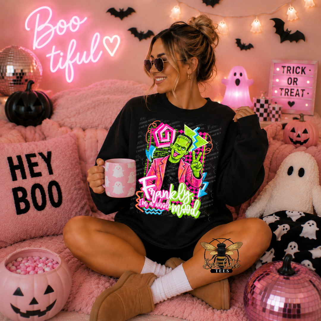 A woman sits cross-legged on a pink blanket in a Halloween-themed room, wearing a black graphic sweatshirt with a colorful Frankenstein-themed print and holding a pink mug. The scene includes pumpkins, ghost pillows, a neon sign, and a light box.