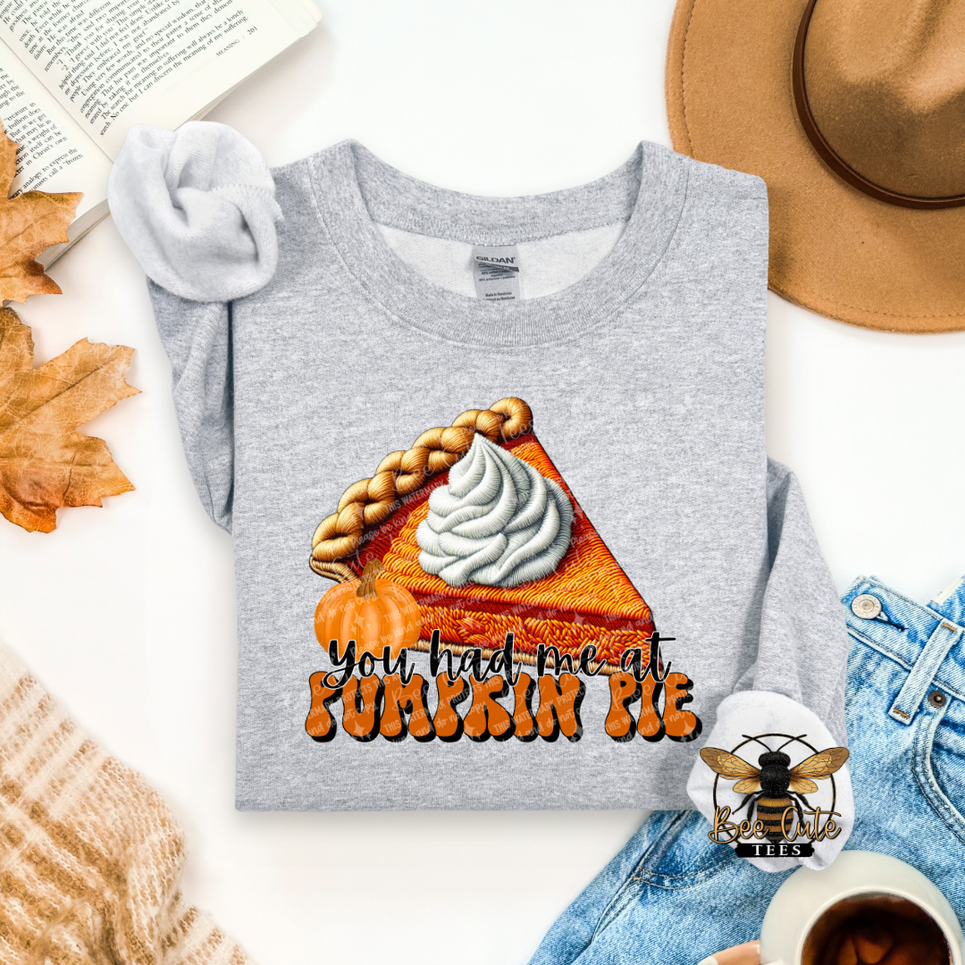A folded heather gray long-sleeve T-shirt with a large autumn-themed pumpkin pie graphic and the text 'You had me at PUMPKIN PIE' in orange and black. The shirt is styled in a flat lay with a tan hat, blue jeans, a book page, fall leaves, a knit sweater, and a cup of coffee on a white background.