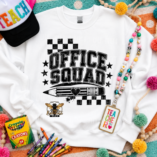 A white crewneck sweatshirt laid flat with a black 'OFFICE SQUAD' graphic featuring stars, a pencil, and a bee. The sweatshirt is styled with a colorful 'TEACH' cap, a beaded necklace, a 'TEACHER' tag, crayons, and a box of Crayola crayons on a patterned background.