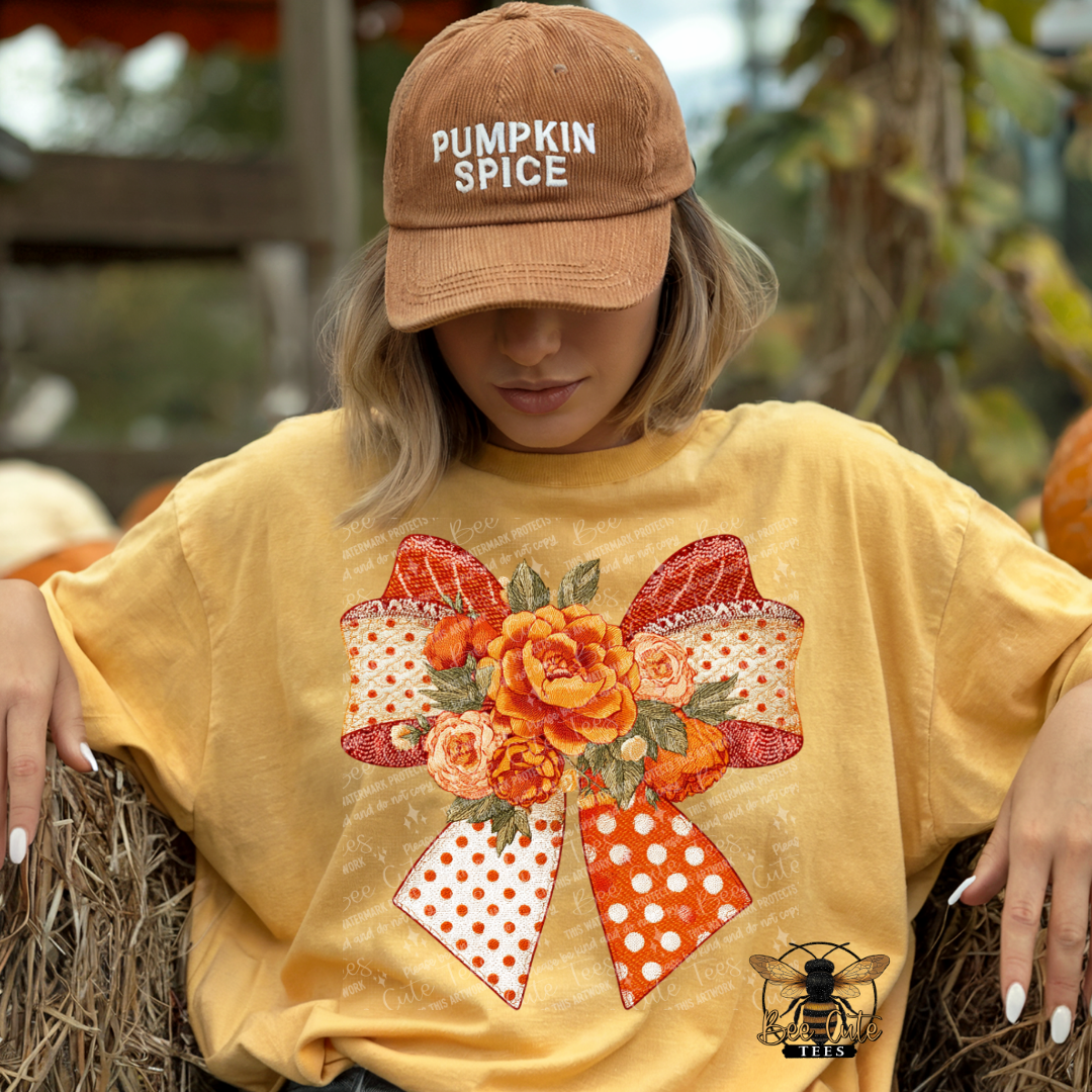 A woman wearing a tan 'Pumpkin Spice' baseball cap and a yellow graphic t-shirt with a large orange and red floral bow design, sitting on a hay bale in an outdoor autumn setting.