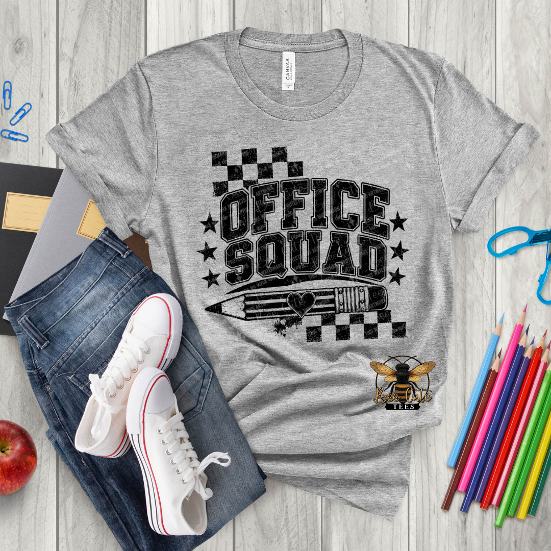 A heather gray graphic T-shirt laid flat on a light wood surface, styled with jeans, white sneakers, a red apple, colored pencils, and paper. The shirt has a large black 'OFFICE SQUAD' graphic with stars, checkered flags, a pencil illustration, and a bee logo near the lower right.
