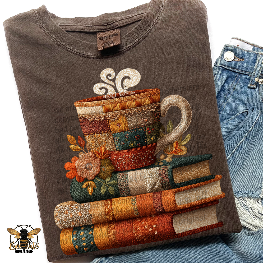 A folded dark brown t-shirt featuring a detailed graphic of stacked books and a mug made from colorful patchwork fabrics, styled next to a pair of blue denim jeans.