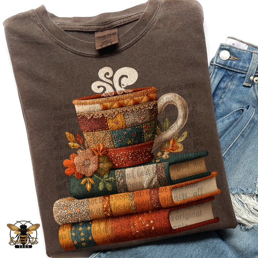 A folded dark brown t-shirt featuring a detailed graphic of stacked books and a mug made from colorful patchwork fabrics, styled next to a pair of blue denim jeans.
