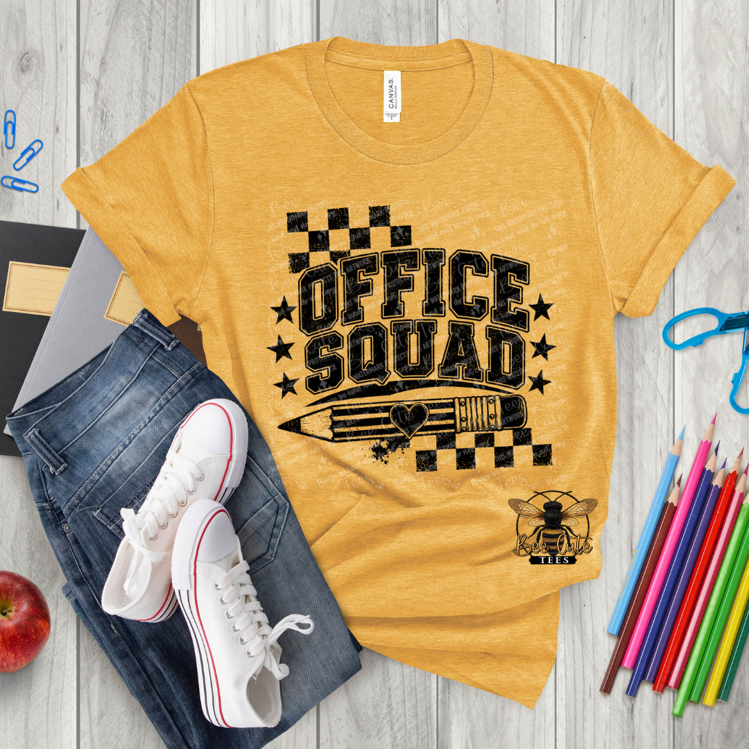 A heathered mustard yellow graphic t-shirt featuring a black 'OFFICE SQUAD' design with stars, a pencil, and a bee logo. The t-shirt is styled in a flat-lay arrangement with blue jeans, white sneakers, colored pencils, a red apple, and a notebook on a gray wood-plank background.