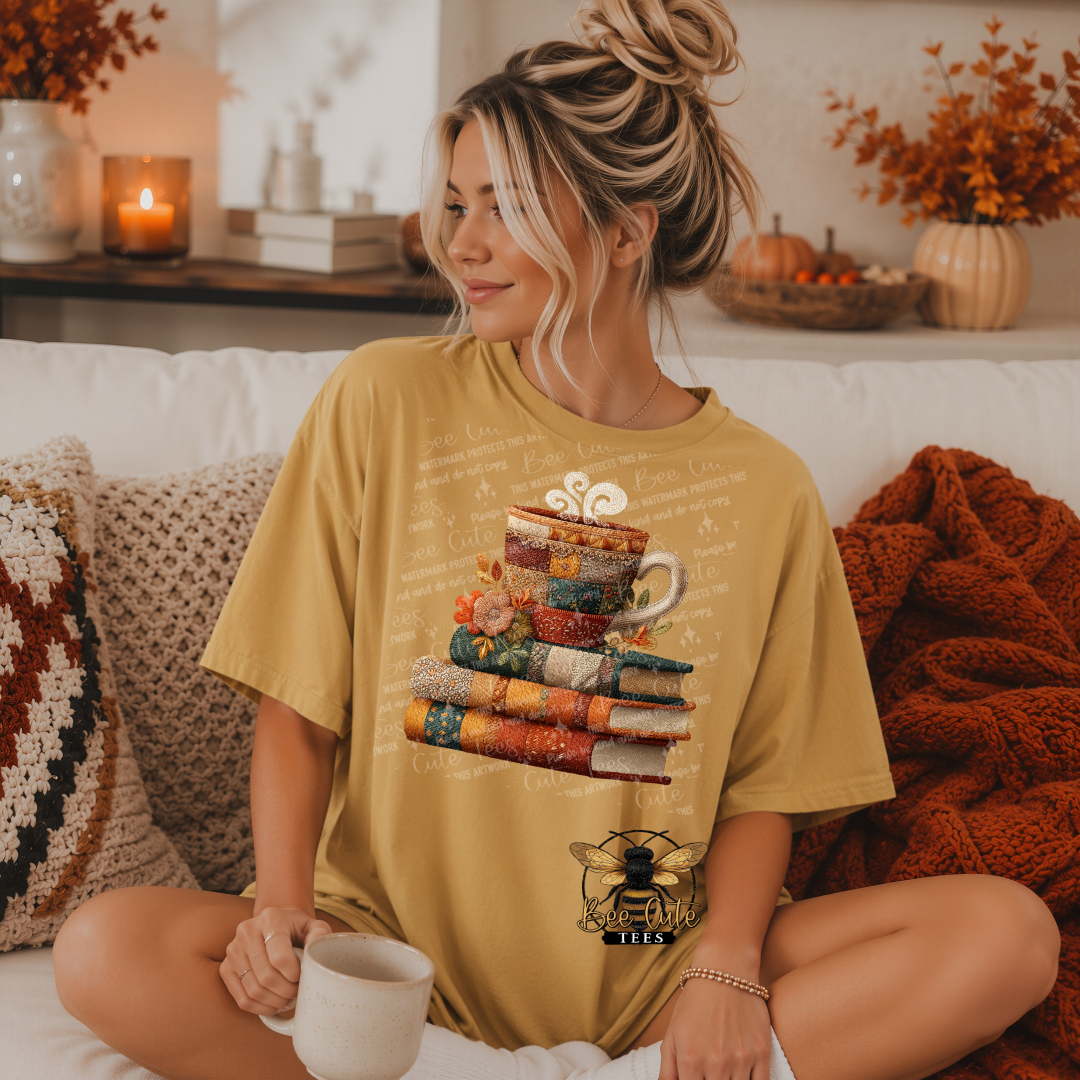 A woman sits cross-legged on a white couch wearing a mustard yellow graphic t-shirt. The shirt features a stack of colorful patterned books with a coffee cup and steam graphic, and a bee logo with the text 'BEE CUTE TEES' at the bottom.